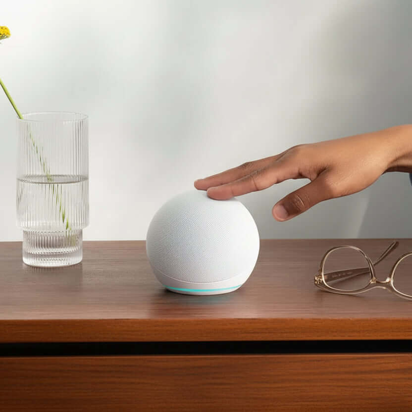 Echo Dot 5th Gen smart speaker in Glacier White on a wooden table next to a glass of water and eyeglasses.