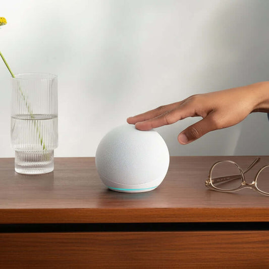 Echo Dot 5th Gen smart speaker in Glacier White on a wooden table next to a glass of water and eyeglasses.